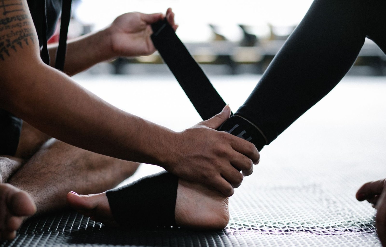A trainer wrapping a black elastic support strap around a bare ankle on a gym mat