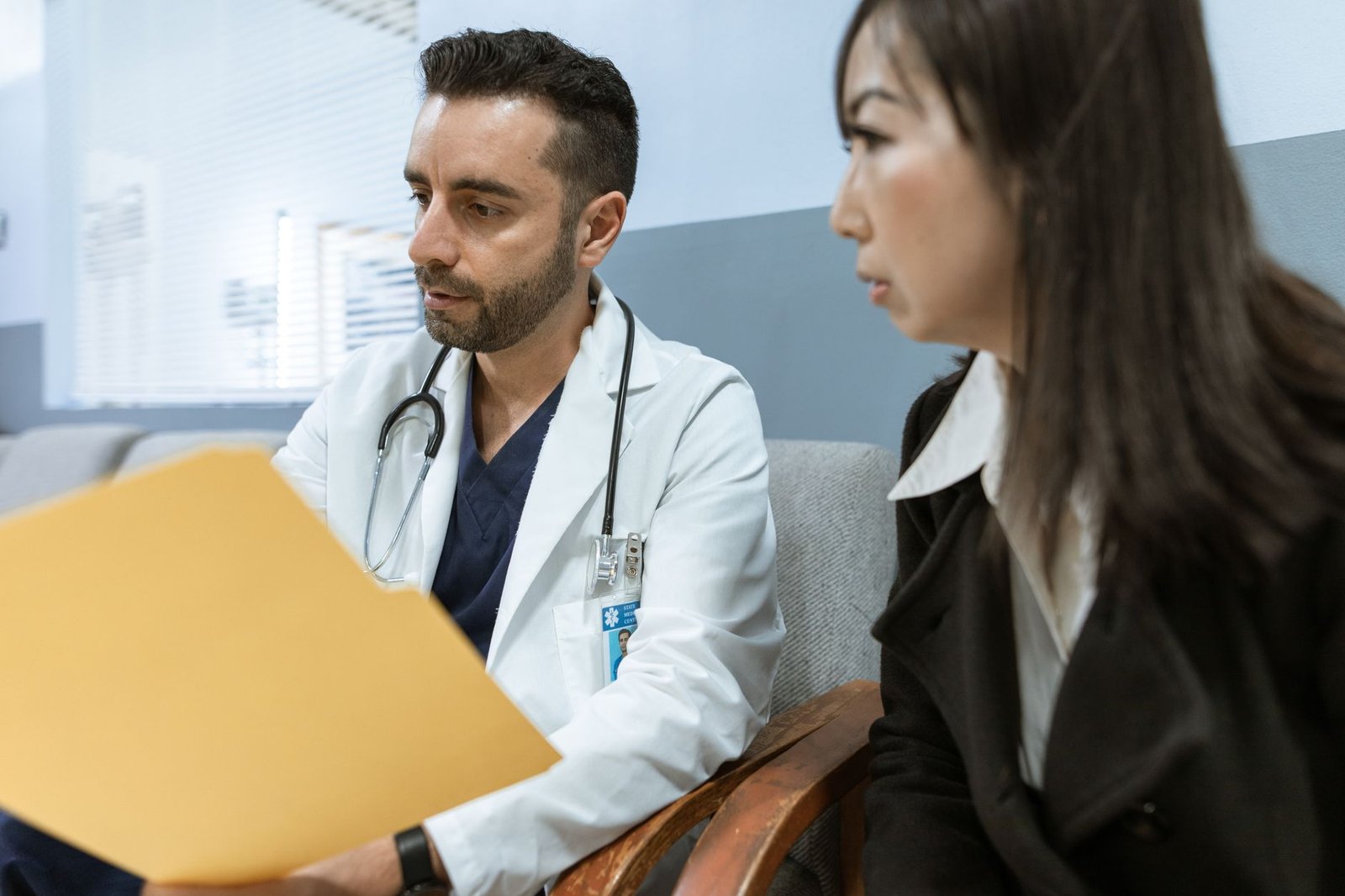 A physician in a white coat reading from a patient file while seated beside the patient during a consultation