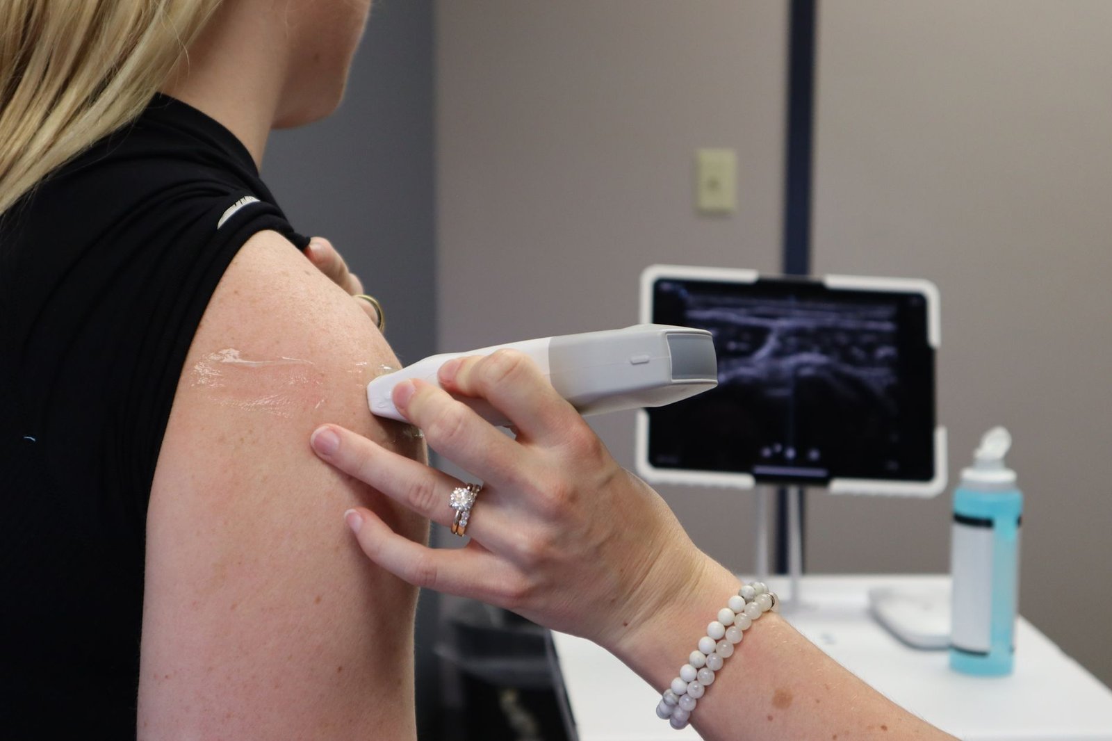 An ultrasound probe held against the front of a shoulder, with the live tendon image on the screen behind