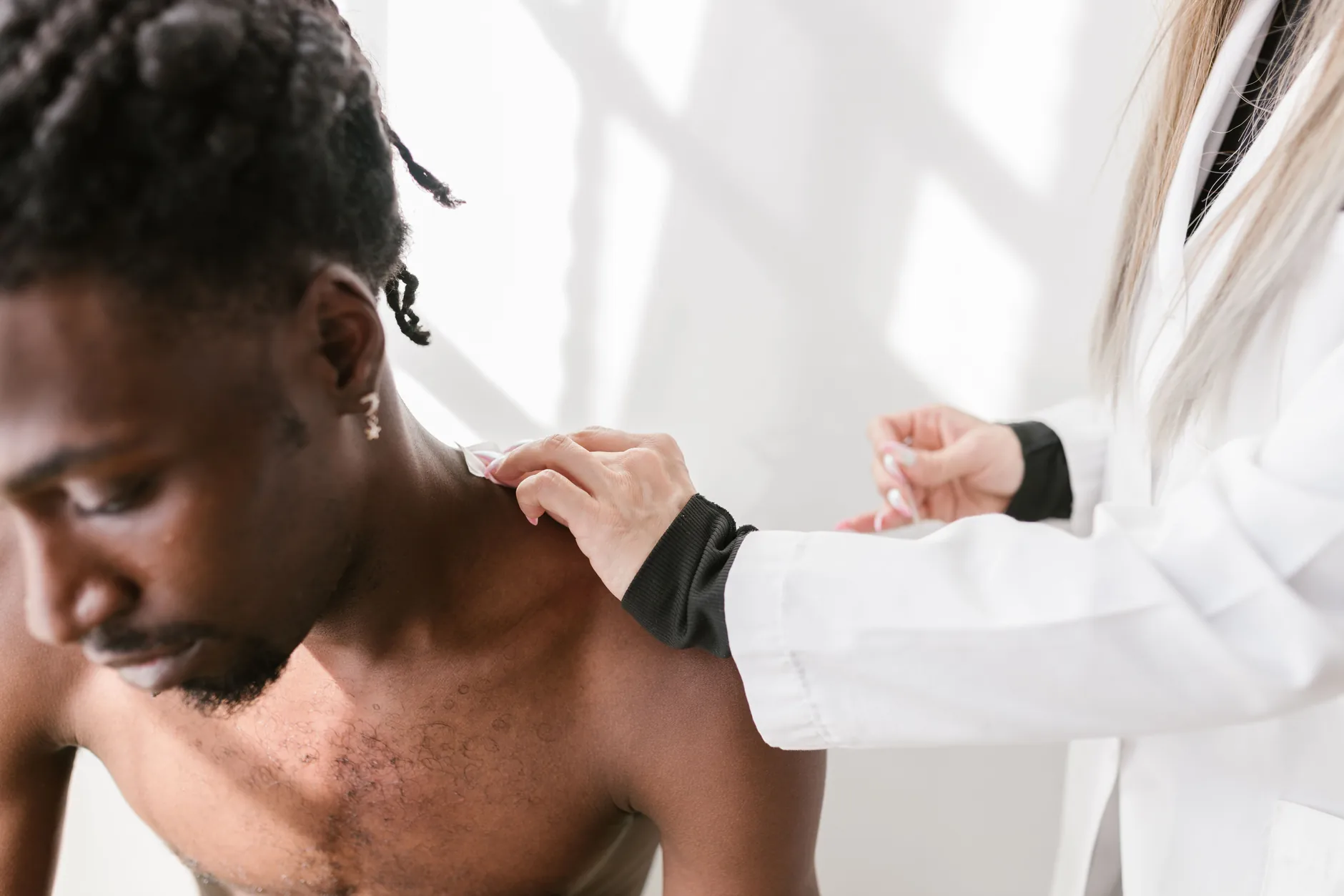 A clinician in a white coat examining a man's painful shoulder with both hands.